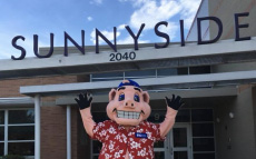 F&M's kid's bank mascot standing in front of a school. 